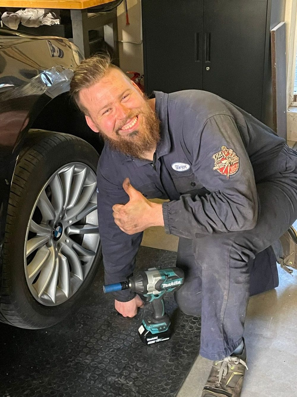 Matt kniet lächelnd mit Daumen hoch neben einem Autorad, Schlagschrauber in der Hand